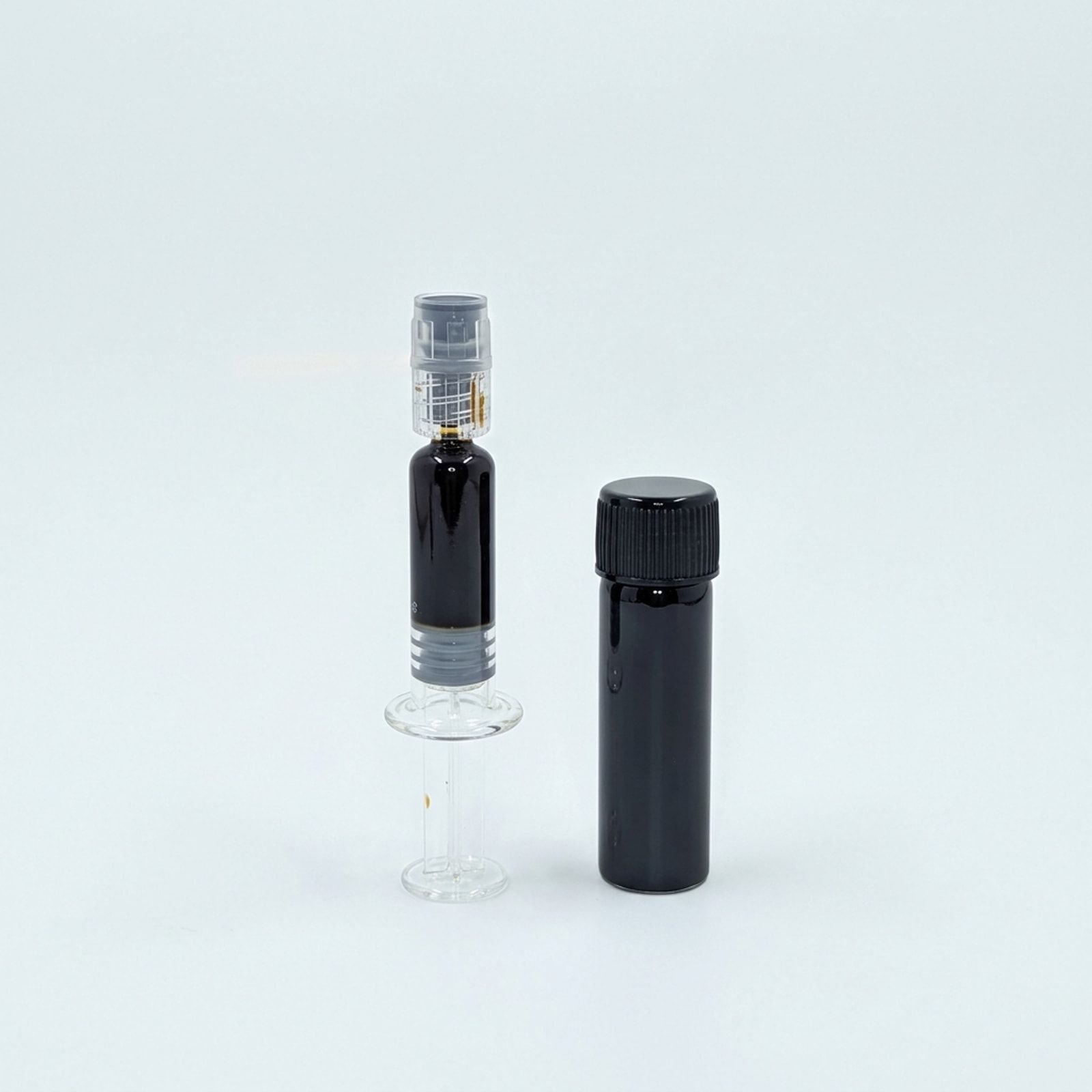 A glass syringe of black oil standing beside a capped black vial, on a white background.