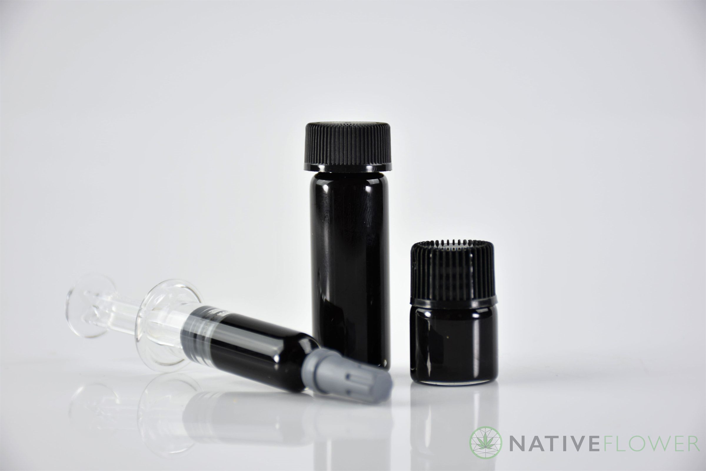 A glass applicator of dark cannabis oil lying beside two small black jars.