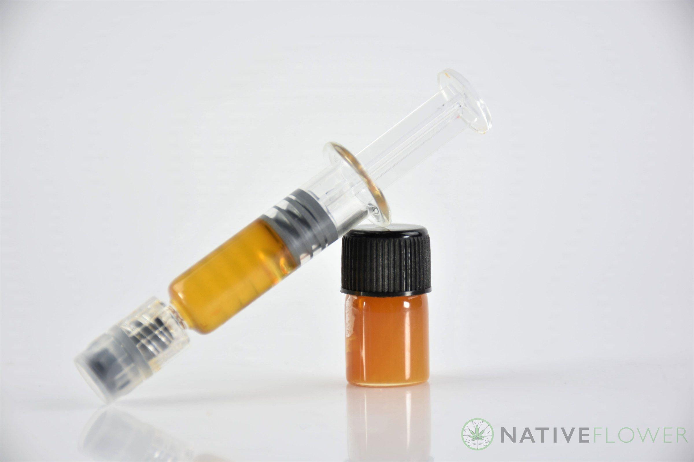 A glass syringe of amber cannabis oil resting against a small vial of the same oil under a black cap.