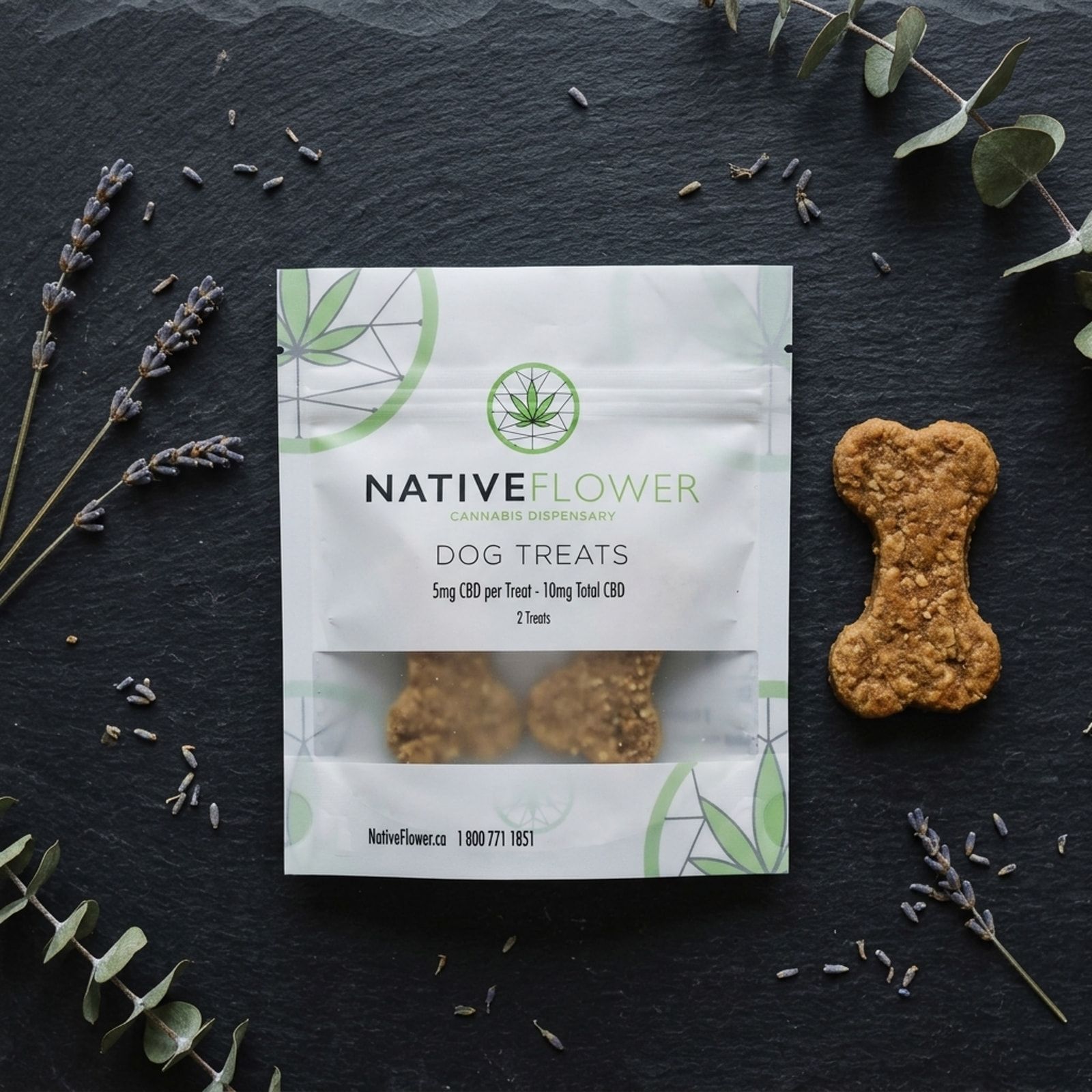 A white Native Flower dog treats pouch on dark slate beside a bone-shaped biscuit, with lavender sprigs and eucalyptus around it.