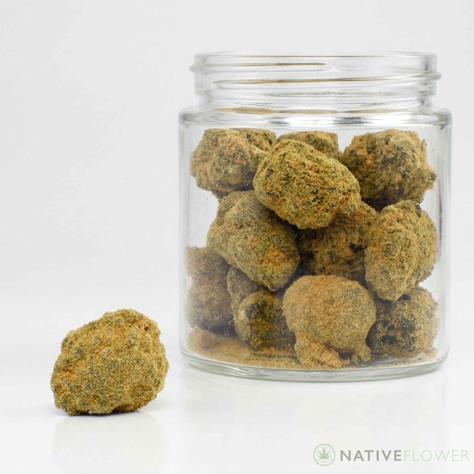 A clear glass jar filled with kief-coated moon rocks, one more resting in front of it, on a white background.