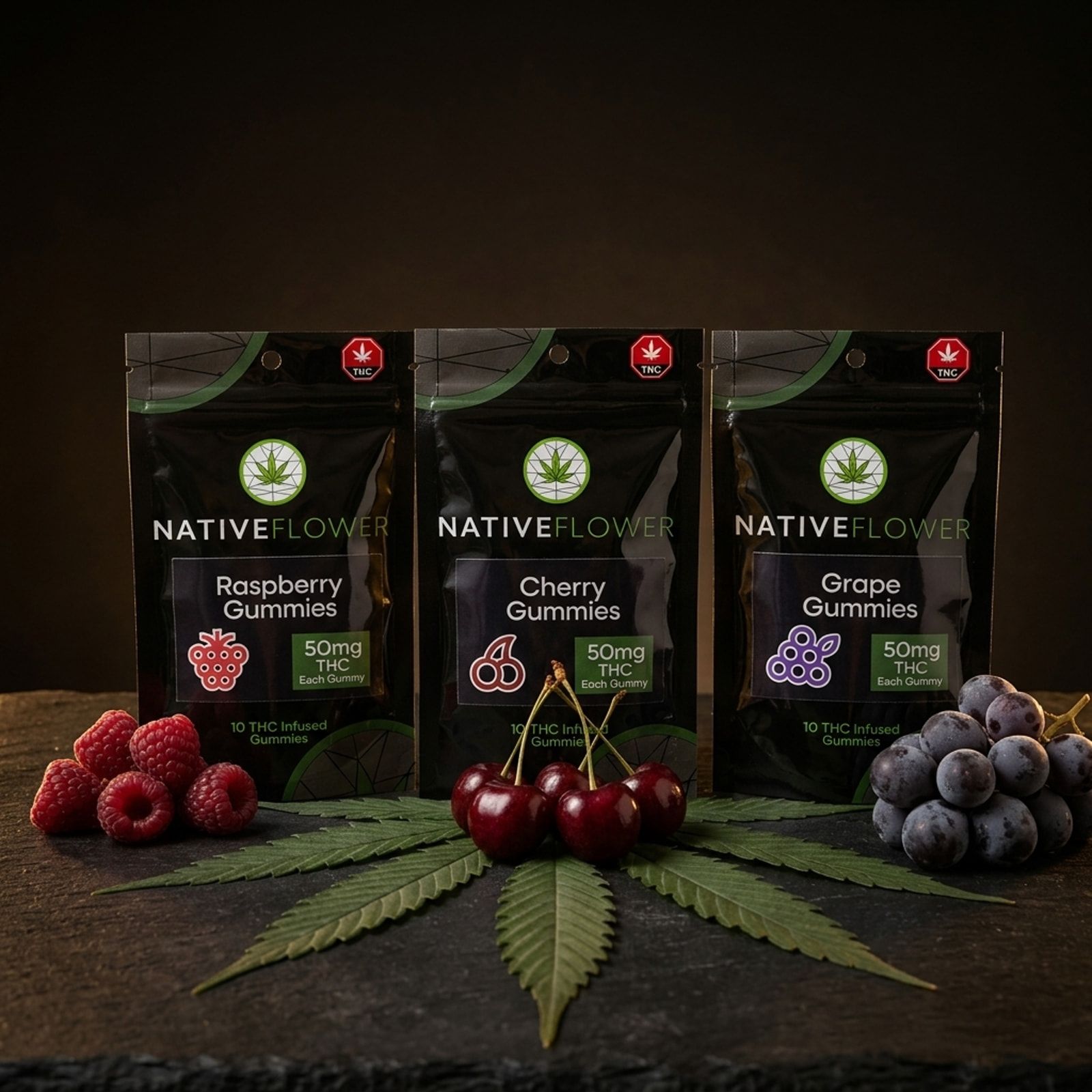 Three black Native Flower gummy pouches — raspberry, cherry and grape — behind fresh fruit arranged on a large cannabis leaf.