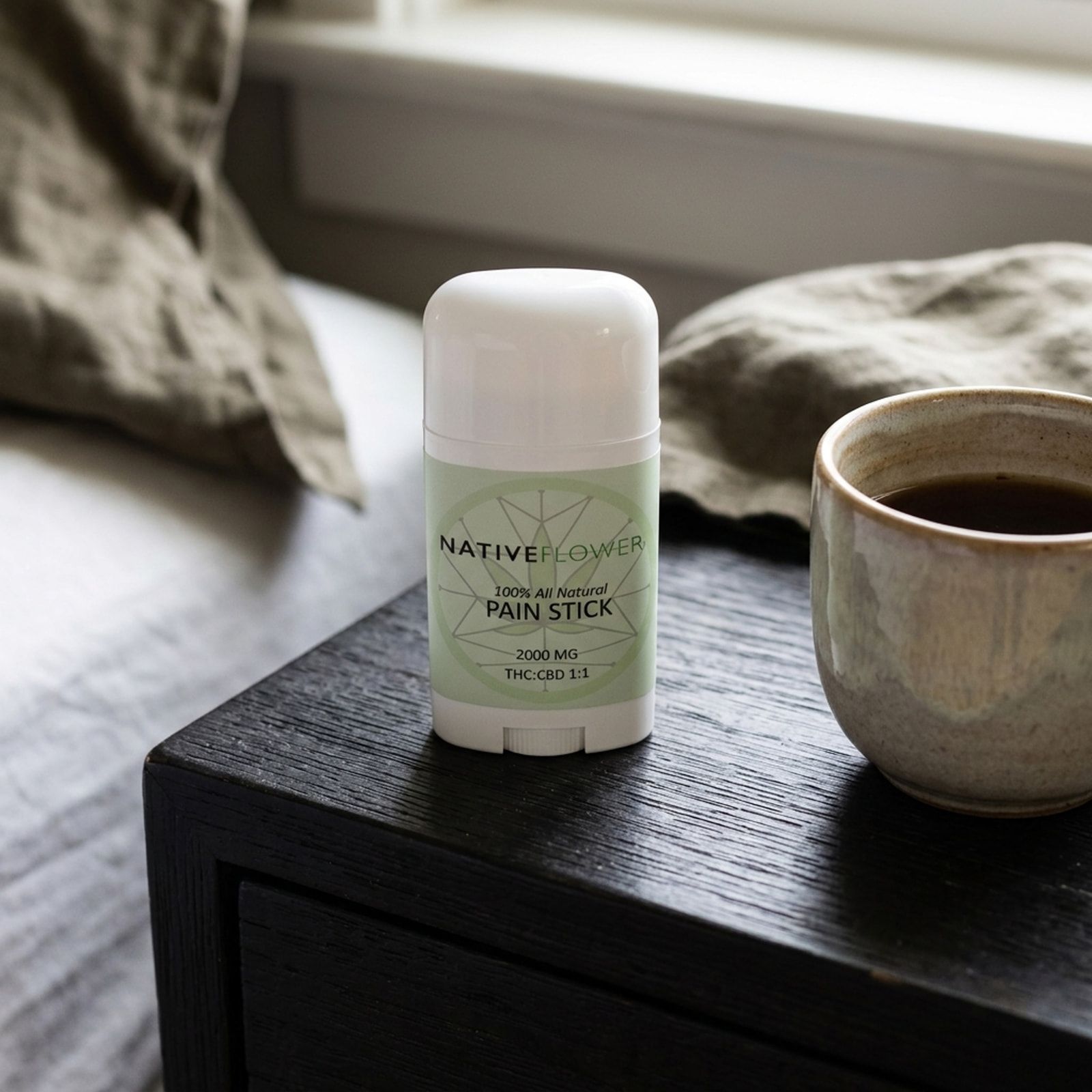 The Native Flower pain stick standing on a black bedside table beside a stoneware cup, rumpled linen bedding behind.