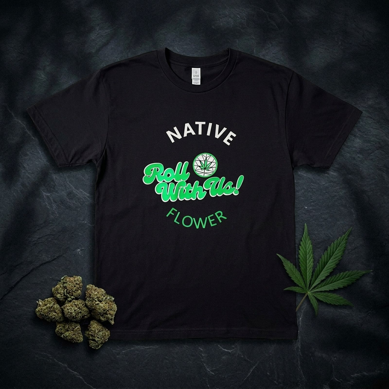 A black Native Flower T-shirt laid flat on dark slate, Roll With Us printed in green across the chest, beside cannabis buds and a leaf.