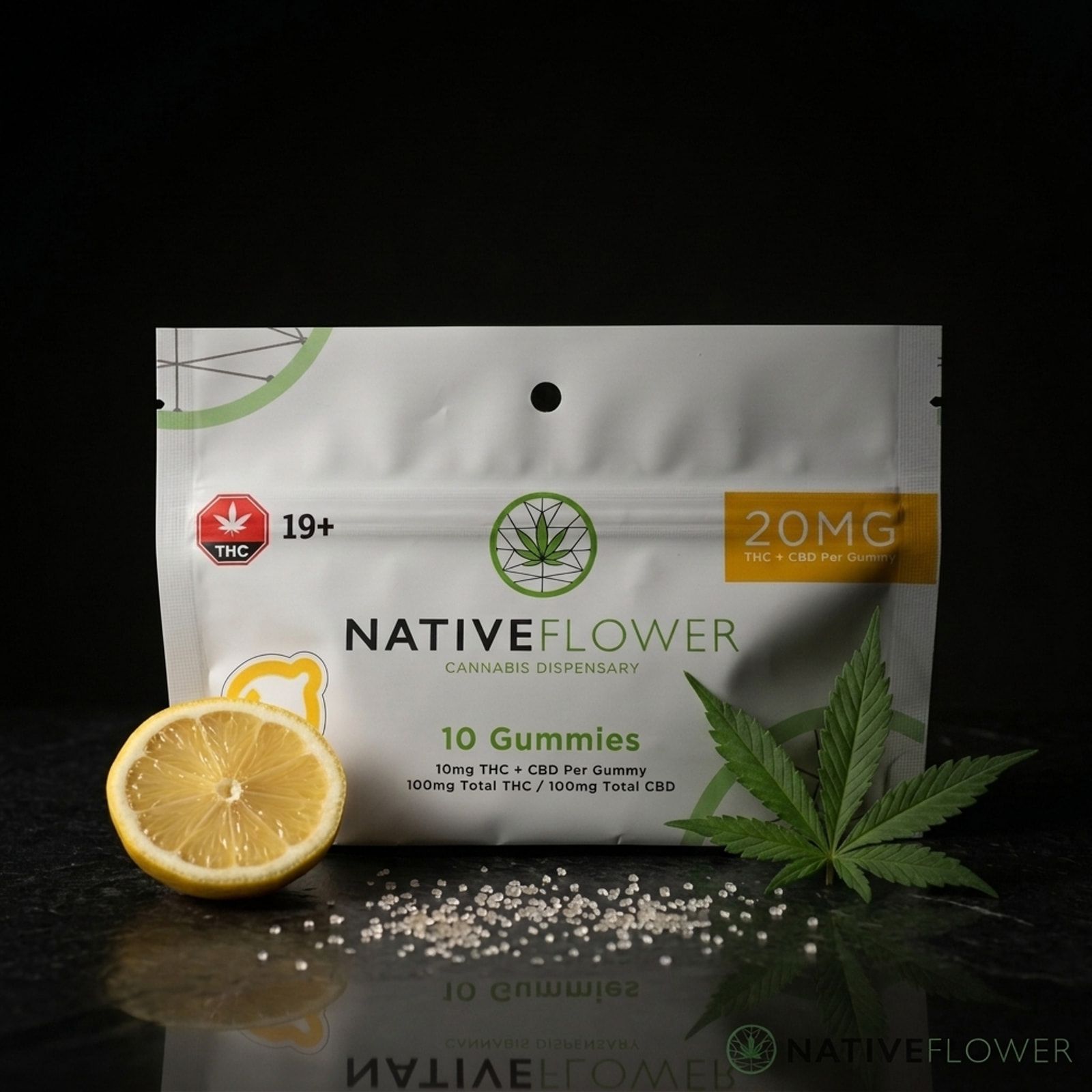 A white Native Flower lemon THC and CBD gummies pouch on a glossy black surface, with half a lemon, sugar crystals and a cannabis leaf.