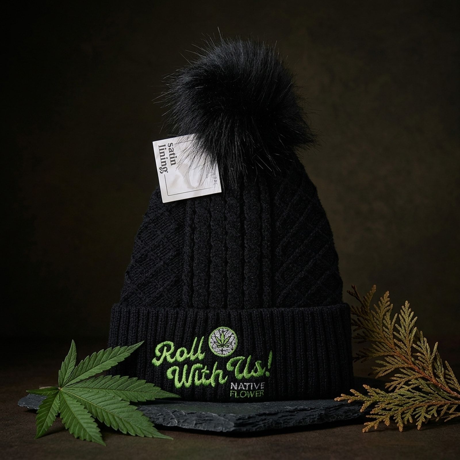 A black cable-knit Native Flower toque with a pom pom on slate, Roll With Us embroidered in green on the cuff, beside a cannabis leaf.