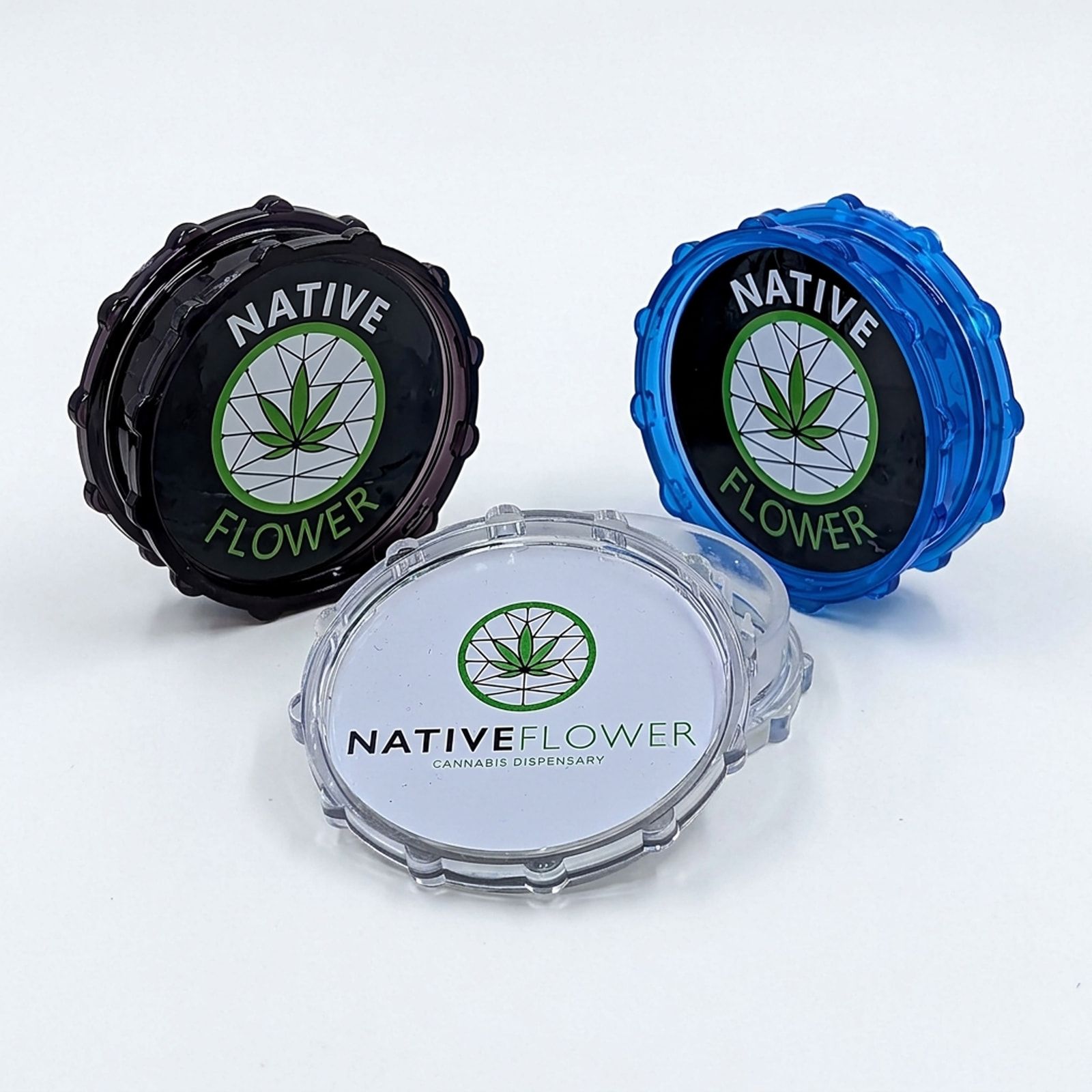Three Native Flower plastic grinders in black, blue and clear on a white background, each lid showing the Native Flower logo.