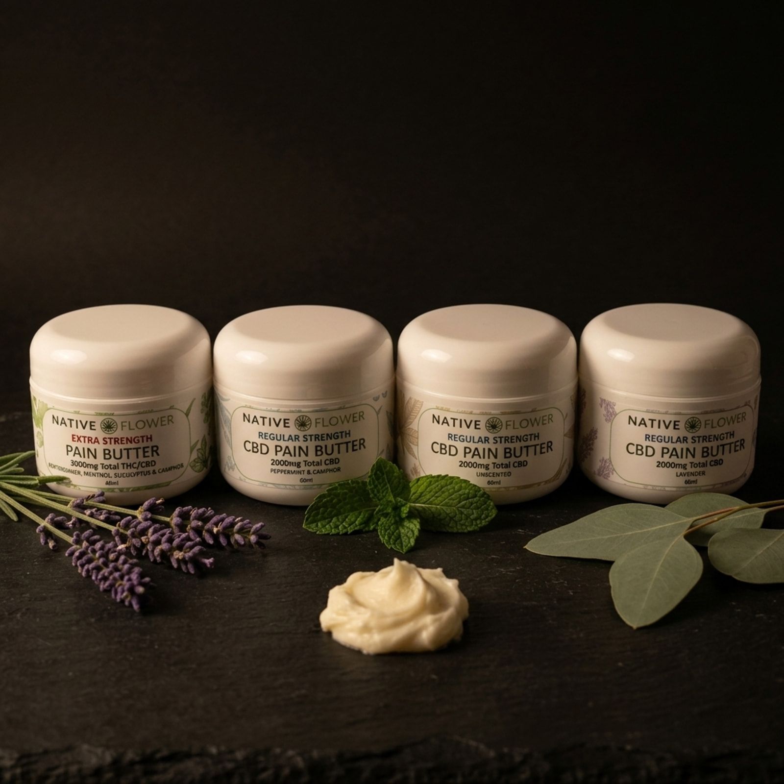 Four white tubs of Native Flower topical butter in a row on slate, with lavender, mint, eucalyptus and a dab of butter in front.