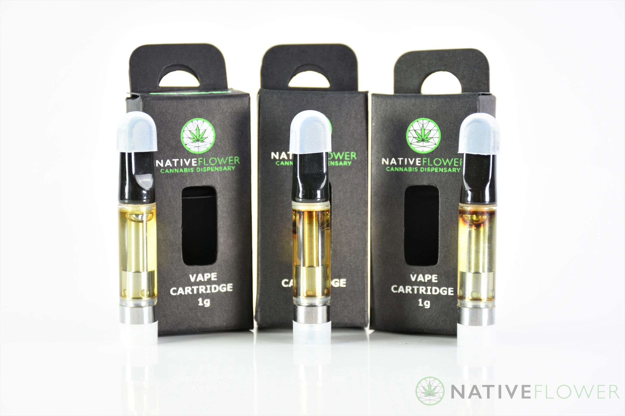 Three glass vape cartridges of golden oil standing in front of their black boxes.